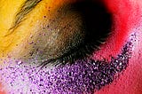 A picture of a closed eye with a painted black eyelid surrounded by dark pink, purple glitter, and yellow around the eyebrow.