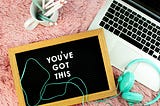 Aerial view of pink plushy carpet. Coffee mug with wooden pencils, an open Macbook, teal over-ear headphones, and a letter board with the phrase, “You’ve got this.”