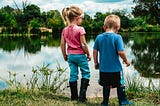Small girl in pink tshirt with blonde ponytail and small boy with short blonde hair and blue tshirt, looking out at the river from the bank.