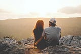 A man and woman are sitting on some rocks looking out over a cliff; the camera is behind them so we see only the edge of their faces. The woman has long hair and is in black. The man is next to her with his arm around her. He is wearing a t-shirt and backwards cap.
