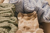 An image of a wicker basket with soft baby blankets in blues and greens, neatly folded next to folded baby clothes.