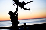 Along a wooden dock fence in the forefront of a distant orange sunset mirroring the dark, thin horizon on the water’s edge are the silhouettes of a Father holding his daughter high in the air with one hand. The daughter’s long hair is scattered around in a web of delight, and her trusting posture reveals how often her Father has lifted her up in the air like this. The joy and connection they share are evident by the level of trust with such a maneuver of grace.