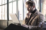 A well-dressed man sits looking at a laptop computer propped on his knees. He appears confused by what he sees.