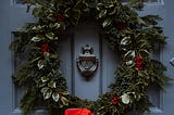 A Christmas garland hangs on a grey front door, circling a metal door knocker