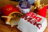 A packaged fast-food meal on a wooden table, with fries, a burger, and a Big Mac box. A familiar, ready-made combination designed for speed, convenience, and immediate satisfaction rather than attention or presence.