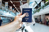person in an airport holds up a blue US passport and a plane ticket