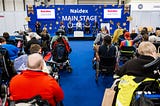 A photograph of the audience and a panel of speakers on the main stage at Naidex. Many of the audience and speakers are wheelchair users.