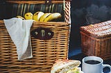 A picnic basket full of banana and a plate of sandwhiches.