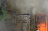 The front of a small old house shrouded in smoke, fire light seen through the window.