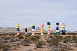 Colorful boulders stacked in seven towers are set against a desert landscape with sparse vegetation and distant mountains under a cloudy sky. These seven towers depict The Seven Components of Authentic A.S.S.™ Infrastructure for coaches, freelancers, and solopreneurs who want to fix their failing systems and achieve business success. Image credit: Lyle Hastie (Unsplash) — “7 rock-based structures in desert field” under the following license: Unsplash Free to Use.