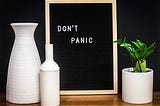 A still life featuring three vases, one containing a plant. There is also a blackboard with the words, Don’t Panic.