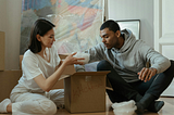 A man and a woman sitting down on the floor while getting things out of a box.