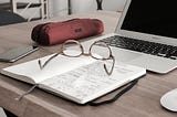 Items are scattered across a wooden desk. Items include a laptop, open notebook, a pair of glasses, and a mobile device.