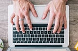 A person’s hands are resting on a keyboard which sits on a wooden desk