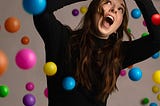 A woman seated looking up hands in the air trying to avoid colorful balls falling on her