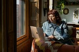 A woman sits on a couch in a homey living room with her laptop in front of her. She is next to a sunny window with sunlight streaming in, appearing calm and focused.