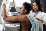 Black women undergoes a mammogram with an Asian women in a white lab coat as the technician.