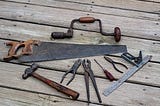 Old tools, their handles brown with age, are arrayed on a weathered wooden floor.