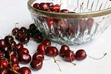 Vintage, crystal bowl with leaded rim, half-full of casually tossed cherries against a white background with more fresh cherries scattered on the white tabletop.