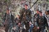 A small group of people in camouflage military uniforms and caps hike down a wooded trail, wearing large backpacks and gear, with relaxed, focused expressions in soft natural light.