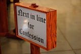 Photograph of a wooden church sign that says “Next in line for Confession” in black text.