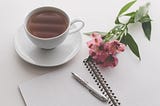 A table with a cup of tea, a pink flower, a notepad and a pen.