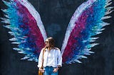 Young woman leaning on a wall with graffiti showing big angelic wings around her.