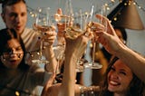 Six people gather around to raise their glasses for a celebration. There are fairy lights and the mood is happy, warm, and fuzzy.