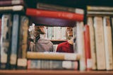 There is a stack of books with a open square in the middle, through it two smiling faces are seen from the nose down. They are facing each other.