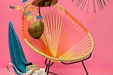 In front of a pink backdrop, an invisible figure, visible only by a hat, shoes and coconut cocktail, reclines on a chair, under a palm tree.
