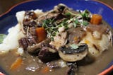 a colorful bowl full of mashed potatoes and covered in homemade coq au vin. Fresh thyme garnishes the dish.
