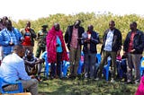 A group of men standing in a group outside with trees in the background. One man wears a traditional red Maasai shuka.