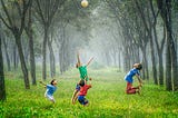 A photo of a four children in brightly coloured shirts jumping up and down on green grass with rows of trees behind them.