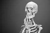 Top half of a skeleton in front of a gray background. Skeleton has one hand in front of its mouth.