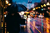 A person with an umbrella on a rainy urban street. The streetlamps are lit.