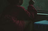 Young child sits inside touching a window with their index finger. The window has drops as it rains outside. The child wears a red jumper and has short hair.