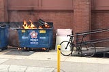 On a city sidewalk is a blue dumpster, bright orange flames peeking out.