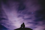 Silhouette of man sitting knees up on hill at night, sky a purple-ish color with clouds