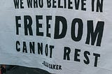 A group of masked people at a protest holding a large banner that reads “We who believe in freedom cannot rest.”