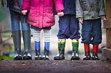 A close-up of four children standing side by side wearing colorful rain boots covered in mud. The lower half of their bodies is visible, showing different pants, leggings, and coats.