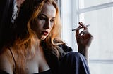 Photo of a light-skinned, copper-haired, distressed young woman wearing a black tank top, smoking a cigarette, in low light by a window.