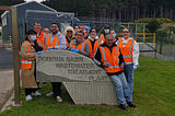 Image description: A group of mana whenua from Ngāti Toa wearing high vis vests are standing around the sign for Porirua Basin Wastewater Treatment Plant. The fence and one of the buildings of the waste treatment plant are in the background.