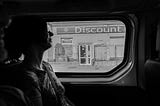 Black and white of a woman sitting in a relaxed position on public transit. Outside the vehicle is a discount store behind some gates.