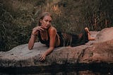 An attractive girl lies sprawled on a rock by some water, woodland in the background.
