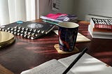 Rowan Ambrose, copywriter, is writing in a notebook with a dark green pencil. The is an empty mug on the dark red desk, with lots of books and notebooks scattered around.
