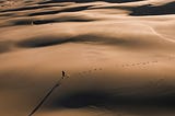 A picture of a person from afar running across a vast desert in the backdrop of a beach in the horizon, and a long line of footprints behind them.