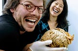 The author holds a brain in the foreground while laughing fiendishly, as Dr. Suzuki laughs from behind.