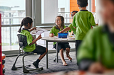 Children in bright green uniform sit and stand around classroom table in New Zealand