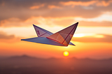 Close-up photo-style image of a paper airplane aloft at sunset with distant mountain range on the horizon
