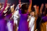 A photo of people dancing with arms in the air in a dark room. There is a lot of motion depicted due to an extended exposure and some figures are brightly lit (white), others less so (purple).